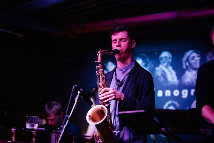 A man wearing a dark blazer and t-shirt plays a saxophone on-stage. There are projections in the background